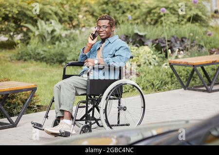 Glücklicher, gutaussehender, behinderter junger afroamerikanischer Mann in einer Brille, der im Rollstuhl sitzt und telefoniert Stockfoto