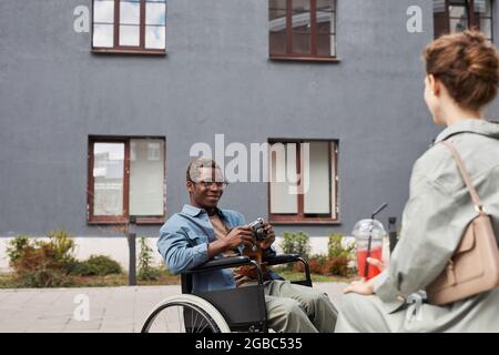 Lächelnder junger behinderter afroamerikanischer Mann in Brille, der im Rollstuhl sitzt und Frau im Freien fotografiert Stockfoto