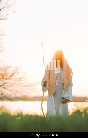 Jesus Christus mit Holzstab steht auf einer Wiese, bekleidet mit seiner traditionellen weißen Robe, vor dem Hintergrund des Flusses und des Sonnenuntergangs. Stockfoto