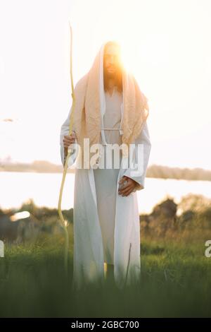 Jesus Christus mit Holzstab steht auf einer Wiese, bekleidet mit seiner traditionellen weißen Robe, vor dem Hintergrund des Flusses und des Sonnenuntergangs. Stockfoto