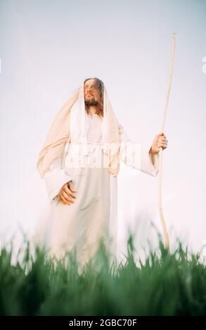 Jesus Christus mit Holzstab steht auf einer Wiese, bekleidet mit seiner traditionellen weißen Robe vor dem Hintergrund des Himmels. Stockfoto