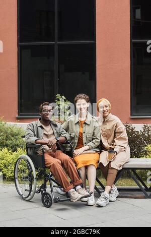 Porträt eines lächelnden jungen behinderten afroamerikanischen Mannes, der im Rollstuhl sitzt und mit multiethnischen Frauen auf der Bank posiert Stockfoto