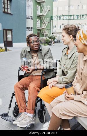 Junger afroamerikanischer behinderter Mann, der im Rollstuhl sitzt und Saft trinkt, während er Zeit mit Freunden auf der Straße verbringt Stockfoto