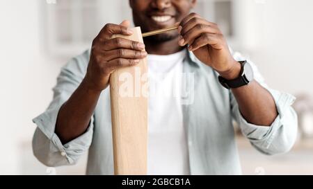 Glücklicher afroamerikanischer Mann, der Schraube an den Holztisch schraubt, den Schreibtisch selbst zu Hause installiert, Nahaufnahme, Ernte Stockfoto