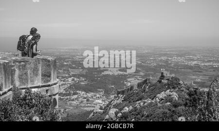 Ein Paar am Rande des Pena Palastes, das von weitem die herrliche Aussicht auf das Schloss der Mauren und die portugiesische Landschaft genießt - Portugal, Sintra - Schwarz Stockfoto