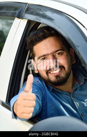 Ein junger indischer Fahrer zeigt den Daumen nach oben und sitzt im Auto Stockfoto