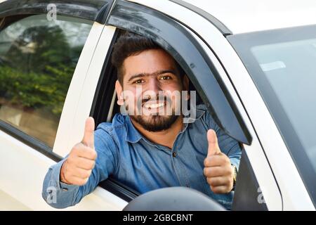 Junger indischer Mann zeigt Daumen nach oben aus dem Auto, Stockfoto