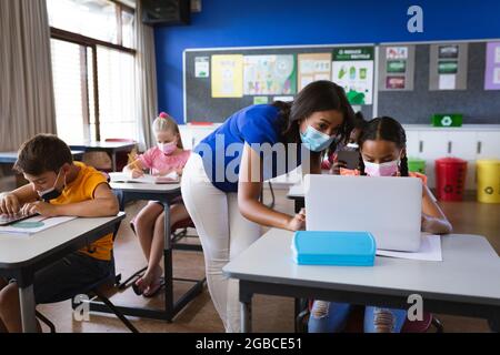 afroamerikanische Lehrerin mit Gesichtsmaske, die einem Mädchen lehrt, einen Laptop in der Grundschule zu benutzen Stockfoto