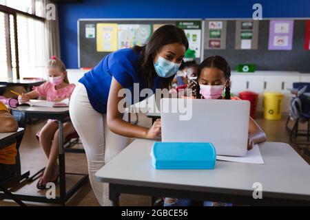 afroamerikanische Lehrerin mit Gesichtsmaske, die einem Mädchen lehrt, einen Laptop in der Grundschule zu benutzen Stockfoto