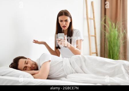 Schockierte Frau, die im Schlafzimmer die Nachrichten des betrügerischen Mannes auf dem Mobiltelefon liest Stockfoto