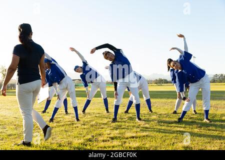 Verschiedene Baseballspielerinnen mit Trainer, die sich auf dem Spielfeld aufwärmen und sich von der Hüfte ausstrecken Stockfoto