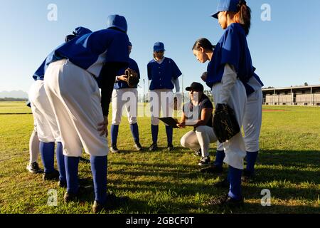 Eine vielfältige Gruppe von Baseballspielerinnen, die auf dem Feld um hockenden Trainer herumstehen Stockfoto
