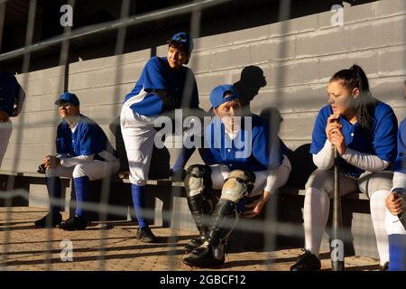 Eine vielfältige Gruppe von Baseballspielerinnen, die in der Sonne auf der Bank sitzen und auf das Spiel warten Stockfoto