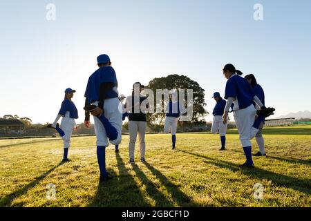 Diverse Baseballspielerinnen mit Trainer, die sich auf dem Spielfeld aufwärmen, stehen, die Beine strecken Stockfoto