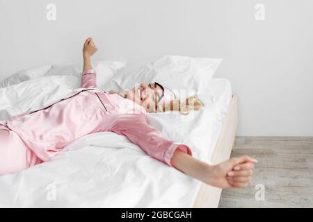 Blonde Frau in rosa Schlafanzug, Schlafmaske aufwachen, streckt Stockfoto