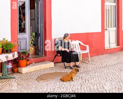 Die alte Frau mit ihrem Hund sitzt an einem warmen Tag vor ihrem Haus. Fuseta Algarve Portugal Stockfoto