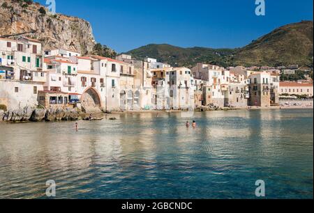 Cefalù, Palermo, Sizilien, Italien. Blick über den ruhigen Hafen auf die Altstadt, überhängende Häuser, die sich entlang der Uferpromenade unter La Rocca gruppieren. Stockfoto