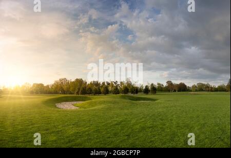 Golfplatz mit Rasen. Abendlandschaft im Park. Blauer Himmel mit Wolken und hellem Sonnenlicht mit Strahlen bei Sonnenuntergang Stockfoto