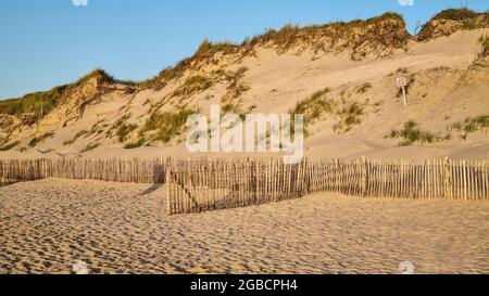 Naturreservat Hügel Sanddünen am Meer durch die Sonne untergehen hinter einem schützenden Holzzaun, bedeckt von Gras. Sauberer Strand. Sign in French: 'Fragile'. Stockfoto
