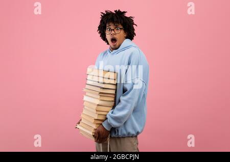 Viele Hausaufgaben. Erschrockener schwarzer Student im Teenageralter, der mit einem riesigen Stapel Bücher stand und den Mund unter Schock öffnete Stockfoto