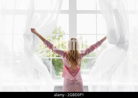 Junge hübsche blonde Frau in rosa Schlafanzug wachte auf, Blick auf große Panoramafenster im Schlafzimmer Stockfoto