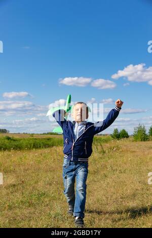 Ein sechsjähriger Vorschulkind in blauer Jacke startet an einem Sommertag ein Spielzeug-Flugzeug auf einem Feld gegen einen blauen Himmel mit Wolken. Die helle Sonne scheint Stockfoto