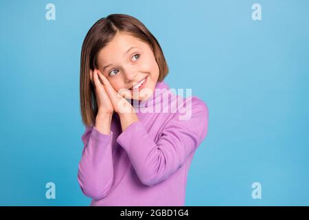 Foto von charmanten hübschen kleinen Schulmädchen gekleidet violett Rollkragen Arme Wangen suchen leeren Raum isoliert blauen Hintergrund Stockfoto