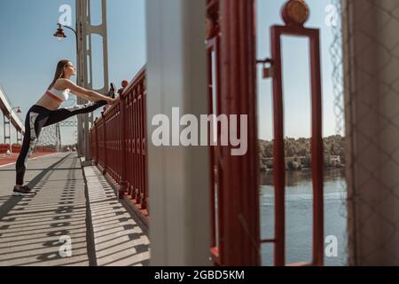 Konzentrierte kurzhaarige Frau macht Dehnungsübungen auf einer zeitgenössischen Brücke Stockfoto