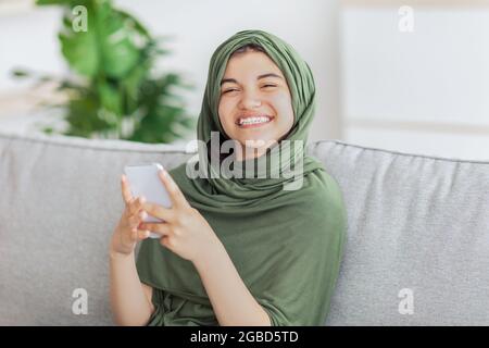 Lächelndes, muslimisches Mädchen im Hijab, das online chattet und zu Hause mit dem Smartphone auf der Couch sitzt Stockfoto