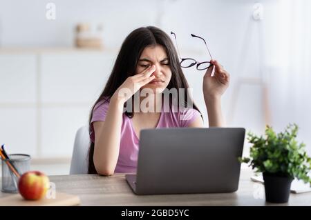 Das indische Teenager-Mädchen fühlt sich müde, reibt ironende Augen, sitzt mit einem Laptop am Schreibtisch, erschöpft von Online-Studien Stockfoto