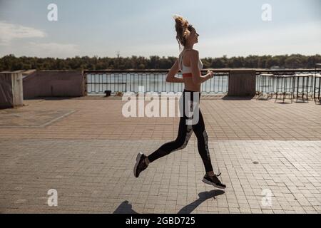 Lady in Sportswear und Sneakers mit lockeren Haaren läuft entlang leerer Stadtwälle Stockfoto