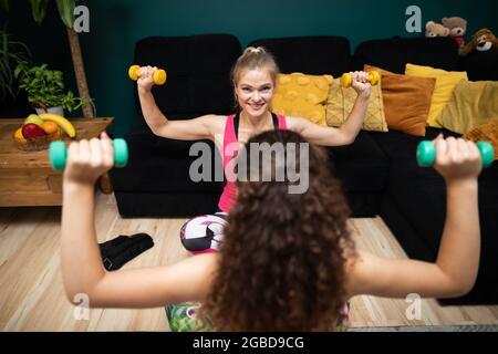 Zwei Mädchen arbeiten, heben ihre Arme mit Kurzhanteln in der Luft. Fitness im Wohnzimmer. Stockfoto