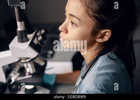 Nachdenkliche asiatische Frau in Uniform mit modernem Mikroskop am Arbeitsplatz im Krankenhauslabor Stockfoto