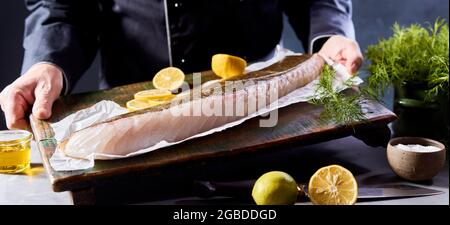 Crop unkenntlich männlicher Koch in schwarzer Uniform demonstriert Holzbrett mit ungekochten Bio-Kabeljau Fischfilet mit Zitronenscheiben während Kochen angeordnet Stockfoto