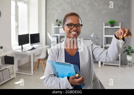 Glücklicher schwarzer Immobilienmakler, der im Büro steht, Schlüssel zum neuen Haus zeigt und lächelt Stockfoto