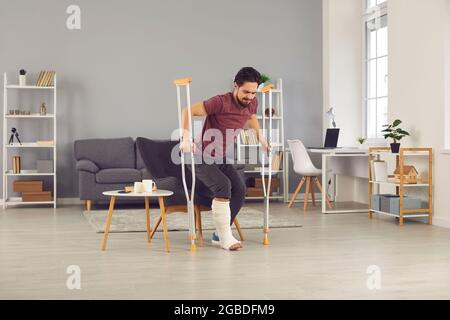 Ein Mann mit gebrochenem Bein versucht, vom Stuhl aufzustehen und mit Krücken im Wohnzimmer zu gehen Stockfoto