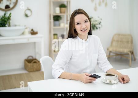 In einem Schönheitssalon trinkt eine junge Frau in einem weißen Hemd Kaffee. Schönes Mädchen Administrator bietet einen Drink an den Besucher. Der Mitarbeiter antwortet Stockfoto