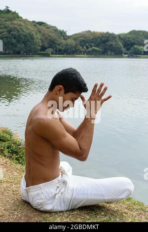 Brasilianischer junger Mann, der am Seeufer sitzt, mit den Händen gegenüber und den Daumen auf der Stirn (Seitenansicht). Stockfoto