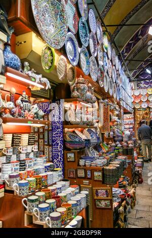 Nahaufnahme der Außenregale eines Souvenirshops mit einer Vielzahl von farbenfrohen Keramiken im Iznik-Stil, darunter Teller, Fliesen, Vasen, Tassen, Und Teekannen, mit einem Mann, der einen gestreiften Pullover und eine Khaki-Hose trägt und im Hintergrund von der Kamera wegblickt, steinerne Fußböden, die ganz rechts zu sehen sind, Und blau geflieste und bemalte Bögen über dem Kopf, die sich in einem gewölbten Durchgang innerhalb des Großen Basars, einem großen überdachten Einkaufsviertel, das von den Osmanen zwischen dem 15. Und 18. Jahrhundert im Fatih-Viertel von Istanbul, Türkei, errichtet wurde, 11. November 2017, befinden. (Foto von Smith Coll Stockfoto