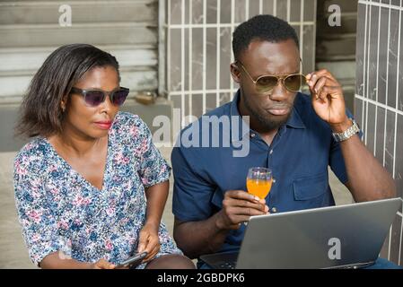 Junge Paare, Frauen und Männer trinken Saft, während sie sich Bilder ansehen oder an einem Laptop arbeiten, der unter einem Gebäude auf der Straße sitzt. Stockfoto