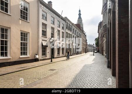 Breda, Niederlande 28. Juni 2021. Eine Straße im Stadtzentrum mit alten hellen historischen Gebäuden, einem Bürgersteig, einer Kirche und einem Radfahrer auf einem bewölkten Stockfoto