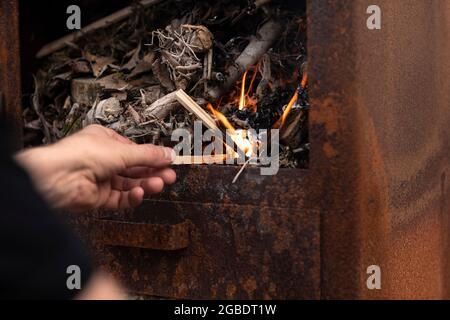 Männliche Hand, die ein Feuer mit kleinen Holzzweigen in einem rostigen Metallofen im Freien macht, während sie sich entspannt und das einfache Leben genießt Stockfoto