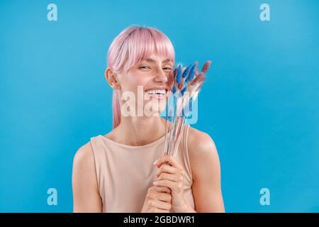 Schöne Frau mit rosa Haaren lächelt der Kamera zu, hält getrocknetes Pampagras in der Hand und posiert über blauem Studiohintergrund Stockfoto