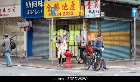 HONGKONG, HONGKONG - 24. Jul 2018: Ein Mann lehnt sich an ein Fahrrad vor einem Immobiliengeschäft an einem roten Hydranten in Hongkong, während eine Dame auf das Fahrrad schaut. Stockfoto