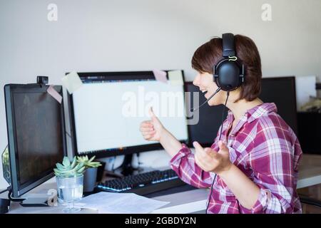 Eine entzückte, emotionale, lächelnde Frau, die auf ihren pc-Bildschirm schaut, Daumen hoch Zeichen tut, während sie am Arbeitsplatz sitzt und einen Videoanruf abruft oder Stockfoto