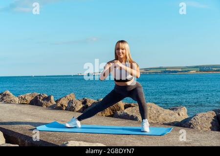 Junge Fitness-Frau im Sport tragen dabei Side-Lunges-Übungen während des Outdoor-Trainings am Strand, Seebrücke. Aktiver Lebensstil und Gesundheitsversorgung Stockfoto