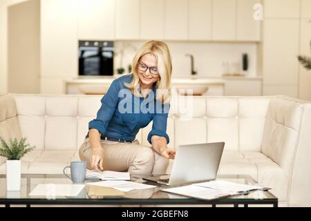 Fröhliche, reife Geschäftsfrau in legerer Kleidung, die Notizen macht, von zu Hause aus arbeitet, während sie auf einer Couch vor dem Laptop in einer modernen Wohnung sitzt Stockfoto