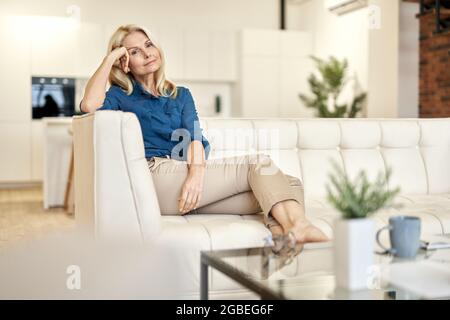 Entspannte ältere Dame in legerer Kleidung, die tagsüber auf einer Couch in einer modernen Wohnung sitzt und die Kamera anschaut Stockfoto