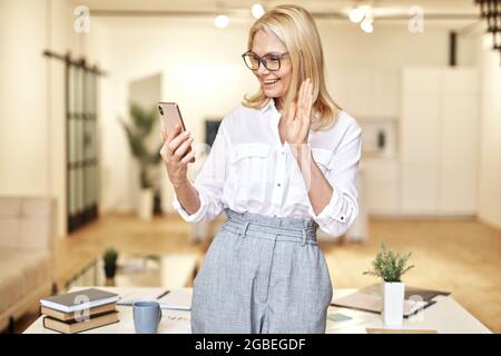 Eine glückliche, reife Geschäftsfrau winkt der Kamera zu und führt Online-Videoanruf mit dem Smartphone, während sie im Büro steht Stockfoto