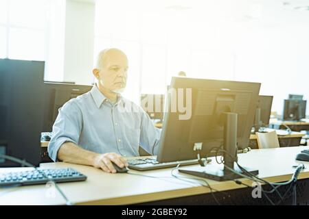 Das Bild eines Geschäftsmannes bei der Arbeit in einem Großraumbüro mit mehreren Arbeitsplätzen, die in seinen Desktop-Computer mit Hintergrundbeleuchtung und strahlendem Sonnenlicht integriert sind Stockfoto